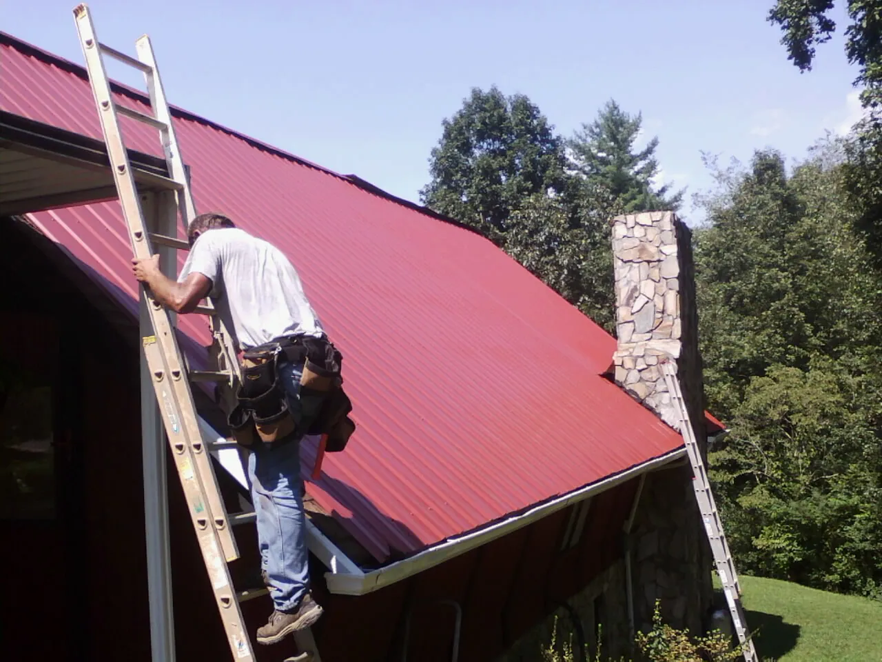 Roofer on a ladder beside a red metal roof and stone chimney — Soffit & Fascia Repair by Roofing Brothers in Whitehouse
